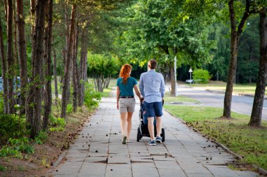 Genç ebeveynler parkta çam ağaçlarının arasında bir bebek arabasıyla yürüyorlar.