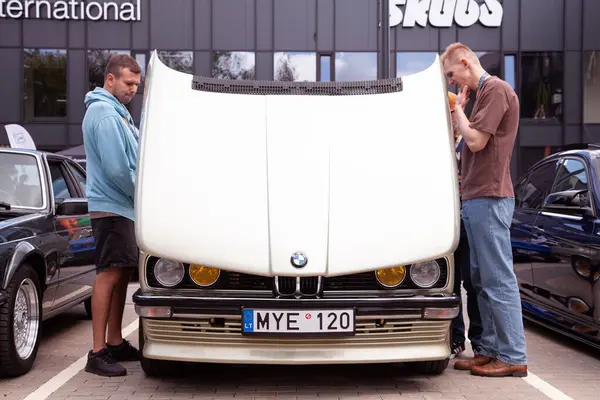 Vilnius, Litvanya - 31-05-2025 - Erkekler, efsanevi BMW 5 serisi E28 'in motor bölmesini bir otoparktaki bir araba karşılaşmasında kaputu açık bir şekilde inceliyor.