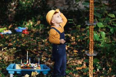 Happy little boy with remote control from radio-controlled toy construction crane in the park in the autumn. Little boy engineer builder architect with safety helmet.
