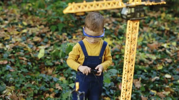 秋に公園で無線制御おもちゃの移動式建設クレーンからのリモートコントロールを持つ小さな男の子の肖像画 安全ガラスの小さな男の子のエンジニアビルダーの建築家 — ストック動画