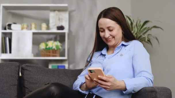 微笑的女孩放松地坐在沙发上 用手机发短信和朋友聊天 快乐的年轻女性在沙发上在线开设智能手机店 或查看手机软件或社交媒体 — 图库视频影像