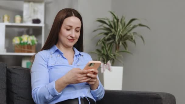 幸せな女性は居心地の良いグレーのソファに休み 現代の携帯電話でインターネットサーフィン スマホを手に自宅で自由な時間を楽しむ白人女性 — ストック動画