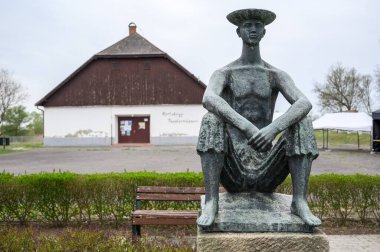 HORTOBAGY, HUNGARY 23, 2022 Bronz bir çoban çocuğu heykeli parkta oturuyor, arka planda kırsal bir bina var. Heykel detaylandırılmış, oturan şapkalı bir çocuğu gösteriyor.