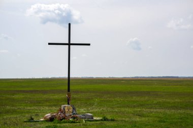 HORTOBAGY, HUNGARY 23 Nisan 2022 Kara metal haçı çoğunlukla bulutlu bir gökyüzünün altındaki yeşil bir alanda duruyor. Haç, küçük ve renkli eşyalarla süslenmiş küçük bir kaya yığınının üzerinde yer alıyor.