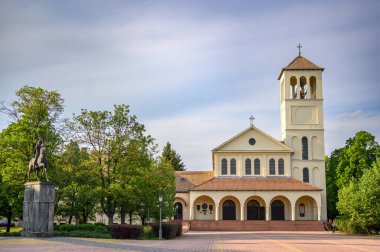 HODMEZOVASARHELY, HUNGARY MAYIS 06, 2022 Sakin bir açık hava sahnesi Bir çan kulesi ile görkemli krem renkli kilise, parçalı bulutlu bir gökyüzü altında yemyeşil ağaçlar ile çevrili. Bir heykel.