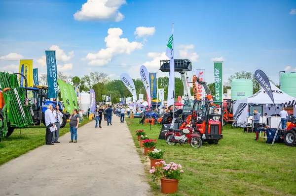 HODMEZOVASARHELY, HUNGARHY MAYIS 06, 2022 Tarım Makineleri Sergisi güneşli bir günde. İnsanlar traktörler ve çiftlik malzemeleri vitrinleri arasında yürüyorlar. Yeşil tarlalar ve mavi gökyüzü