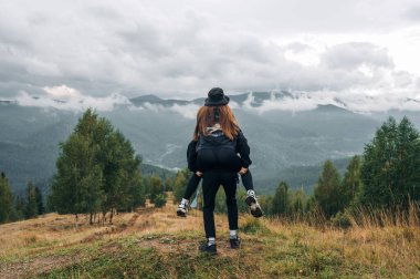 Dağlarda neşeli iki turist, adam kızı sırtında taşıyor, arka planda güzel manzaralar..