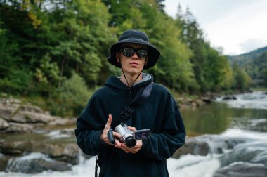 Sıradan giysiler ve panama giymiş genç bir hippi elinde retro kamerayla bir dağ nehrinin arka planında kameraya poz veriyor..