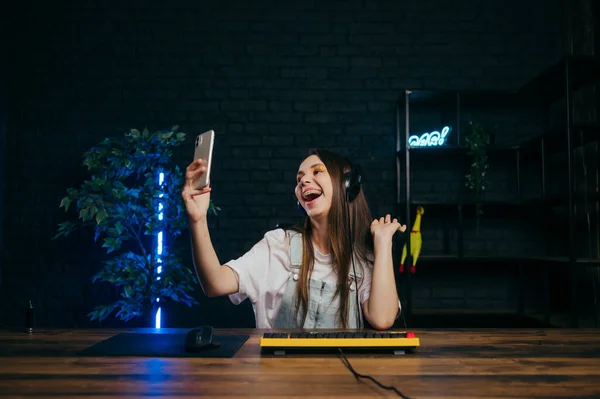 A positive gamer woman takes a selfie while streaming an online game on the computer, smiles and looks into the smartphone camera.