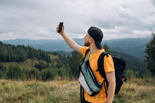 A tourist in casual clothes stands in the mountains and uses a mobile network to view the hiking route, wears a backpack and a cap. Active recreation.