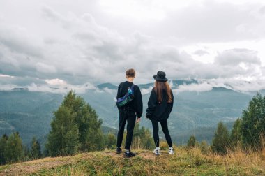 Bir çift turist koyu renk elbiseli kadın ve erkek yürüyüş sırasında dağlarda durup manzaranın güzelliğine ve manzarasına bakıyorlar..