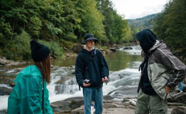 Bir grup turist, dağlarda hızlı kayalık bir nehrin arka planında duruyorlar ve yüzlerinde gülümsemeyle konuşuyorlar..