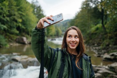 Yürüyüşte bir dağ nehrinin arka planına selfie çeken pozitif bir kadın yürüyüşçü yüzünde bir gülümsemeyle..