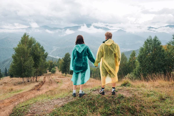 stock image Woman and man tourists in raincoats stand in the mountains and look ahead at cloudy mountain scenery, back view.