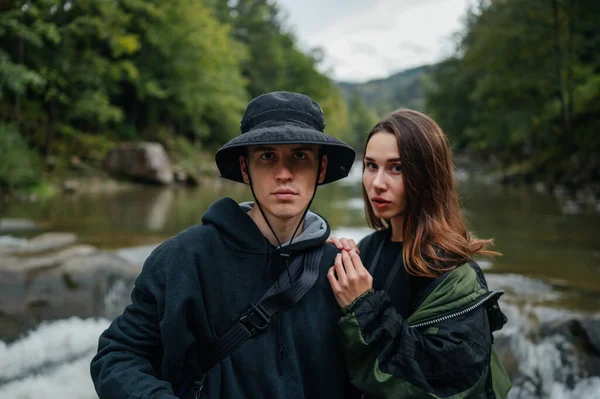 Stylish young couple of tourists on a hike posing for the camera against the background of a mountain river and forest, looking at the camera with a serious face.
