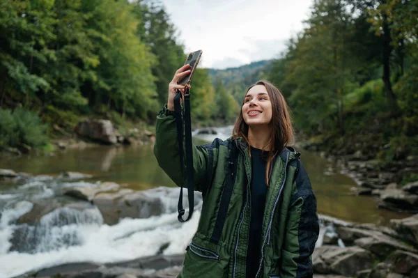 Happy female hiker taking selfie on smartphone against mountain river background while hiking and smiling. Mountain tourism.