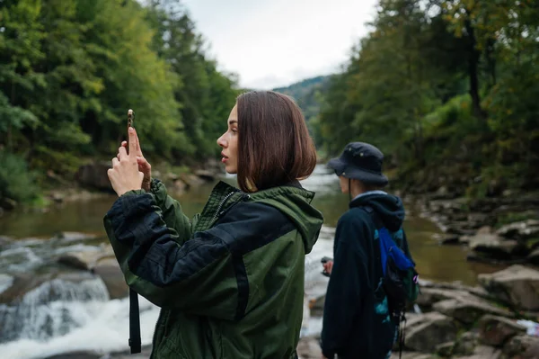 在森林的背景下 一对游客站在一条山河边 一个女人在一个男人的背景下 用智能手机相机拍照 徒步旅行的年轻游客 — 图库照片
