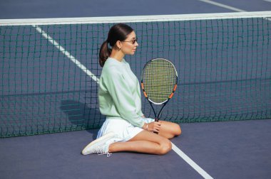 Sportif bayan tenisçi elinde raketle sahanın yanında oturur ve kameraya poz verir, gözünü kaçırır. Tenis oynadıktan sonra dinleniyor.