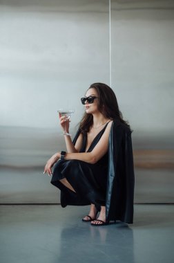 Ceketli ve siyah güneş gözlüklü şık bir kadının portresi. Gümüş duvardaki bir bardaktan alkollü bir içecek içiyor. Profil fotoğrafı..