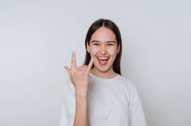 Genç Asyalı kadın gülümsüyor ve açık gri arka plana karşı günlük kıyafetlerle el hareketi yapıyor.