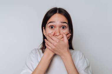 Elleri tarafsız bir ortamda ağzını kapatırken şaşkınlığını ve şaşkınlığını ifade eden bir kadın.