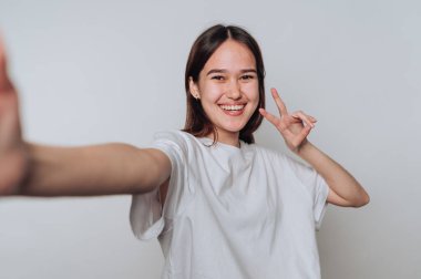 Gülümseyen genç bir kadın, parlak bir stüdyoda barış işareti yaparken selfie için poz veriyor.