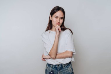 Genç Asyalı kadın, bir eli çenesinde basit bir stüdyo ortamında düşünceli bir şekilde poz veriyor.