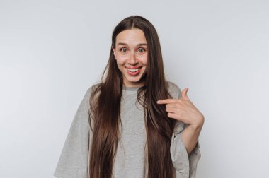 Genç bir kadın, sade bir arka plana şakayla poz veriyor, uzun saçlarını ve neşeli ifadesini göstererek sıradan bir anda