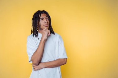 Saçları örgülü düşünceli Afro-Amerikalı genç bir adam parlak sarı bir arka plana karşı poz veriyor, hayatın sorularını yansıtıyor.