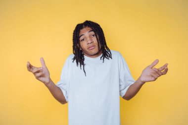 Güneşli bir günde parlak sarı bir arkaplan önünde omuz silkerek kafa karışıklığını ifade eden genç.