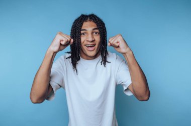 Saçları örülmüş genç bir adam, sıradan bir iç mekan ortamında mavi bir arka plana sevindiğini ifade ediyor.