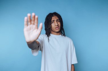 Kıvırcık saçlı genç Afrikalı bir adam mavi arka plana el hareketi göstererek dur ya da dur mesajı veriyor.