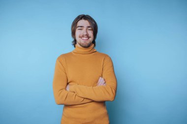 Genç adam, gündüz saatlerinde mavi bir arkaplanda kollarını kavuşturarak güvenle gülümsüyor.