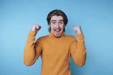 Mavi arka plan karşısında yumruklarını kaldırarak kutlama yapan genç adam, sıradan bir kıyafetle neşe ve heyecan sergiliyor.