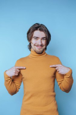 Sarı balıkçı yaka kazaklı genç adam sevinçle mavi arka planda poz veriyor. Pozitif bir tavır sergiliyor.