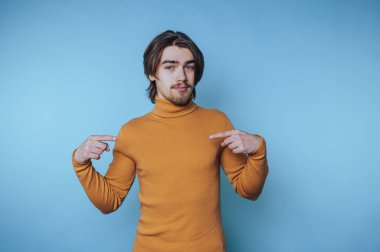 Balıkçı yaka kazak giyen genç bir adam, sıradan bir anda kendisine işaret ederken mavi bir arka plana karşı güvenle poz verir.