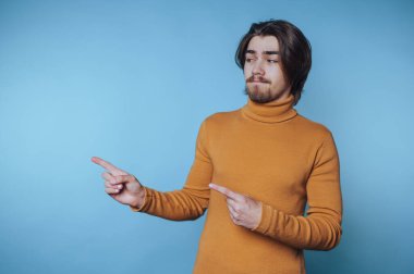 Balıkçı yaka kazaklı genç adam, parmaklarıyla kapalı bir alanda mavi bir zemin üzerinde el kol hareketi yapıyor.