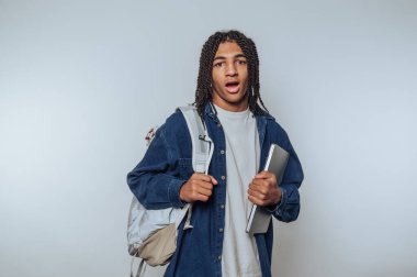 Sırt çantası ve dizüstü bilgisayarı olan öğrenci ev ortamında şaşkınlığını dile getiriyor