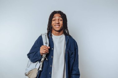 Saçları örgülü genç adam gülümsüyor ve sırt çantasıyla arka planda duruyor.