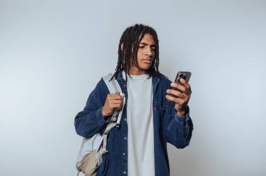 Genç adam akıllı telefonu kontrol ediyor. Sırt çantasıyla kapalı bir alanda rahat bir şekilde duruyor.