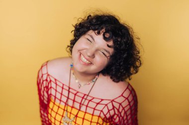 Renkli kıyafetli gülümseyen genç kadın, neşeli ifadeyi kucaklarken parlak sarı arka plana karşı poz veriyor.