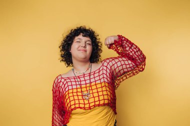 Kendine güvenen genç bir kadın, renkli bir kıyafet ve neşeli bir ifade takınırken canlı sarı bir arkaplan karşısında poz verir.