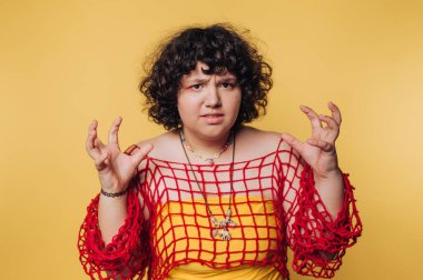 Sarı arka planda kırmızı tüllü bir bluz giyen kadın, ellerini oynak bir şekilde kaldırmış anlamlı bir duruş sergiliyor.