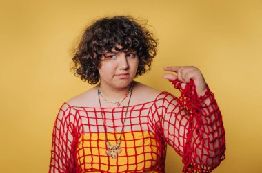 Şahıs, sarı bir elbisenin üzerine kırmızı bir ağ giyip parlak sarı bir arka plana yaslanıyor.