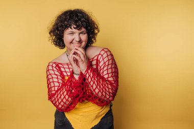 Parlak kırmızı ağlı bluz ve sarı gömlek giyen kişi canlı sarı bir arka plana karşı neşeyle poz verir