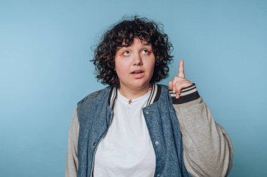 Kıvırcık saçlı genç bir adam mavi bir arkaplan önünde gelişigüzel düşünüyor.