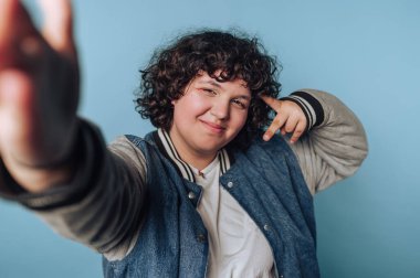 Genç, açık mavi bir arkaplanda güven içinde poz veriyor, barış işareti ile neşeli bir ruh sergiliyor.