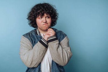 Kıvırcık saçlı çocuk, mavi arka planda düşünceli bir şekilde el ele tutuşurken derin derin düşünür.