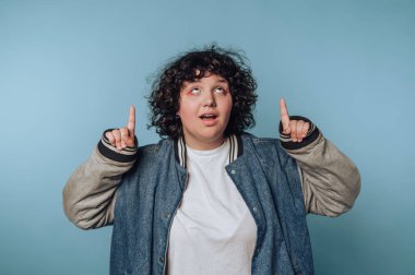 Kıvırcık saçlı genç, gündüz kıyafetleri içinde mavi bir arkaplan üzerinde şakacı bir şekilde yukarı bakıyor.