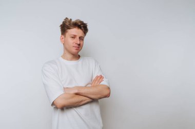 Rahat beyaz tişörtlü genç adam, kollarını çapraz yapıp beyaz bir arka plana doğru poz veriyor.